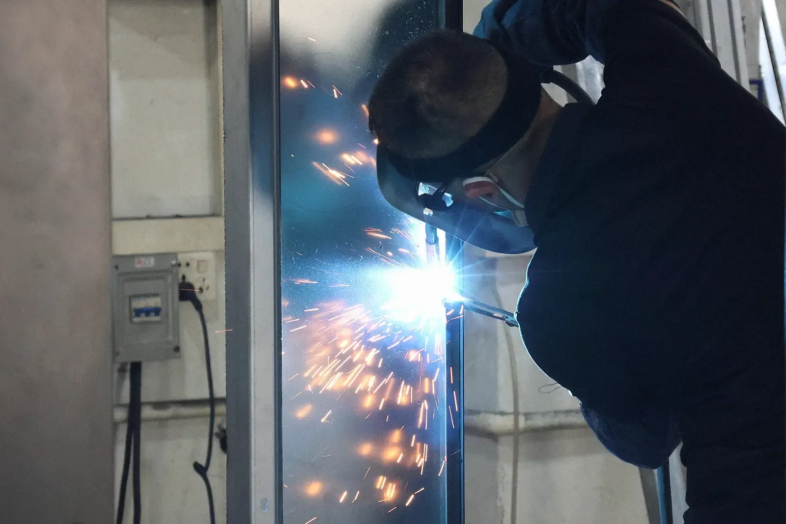 Skilled welder joining steel panels for custom electrical cabinet shells with arc welding in production workshop